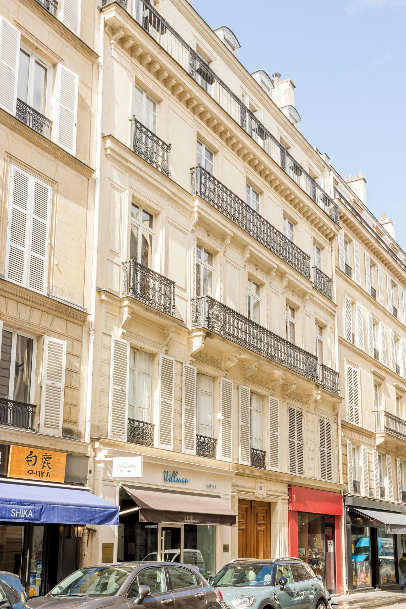 Un immeuble à plusieurs étages, doté de balcons, de devantures de magasins et de voitures garées.