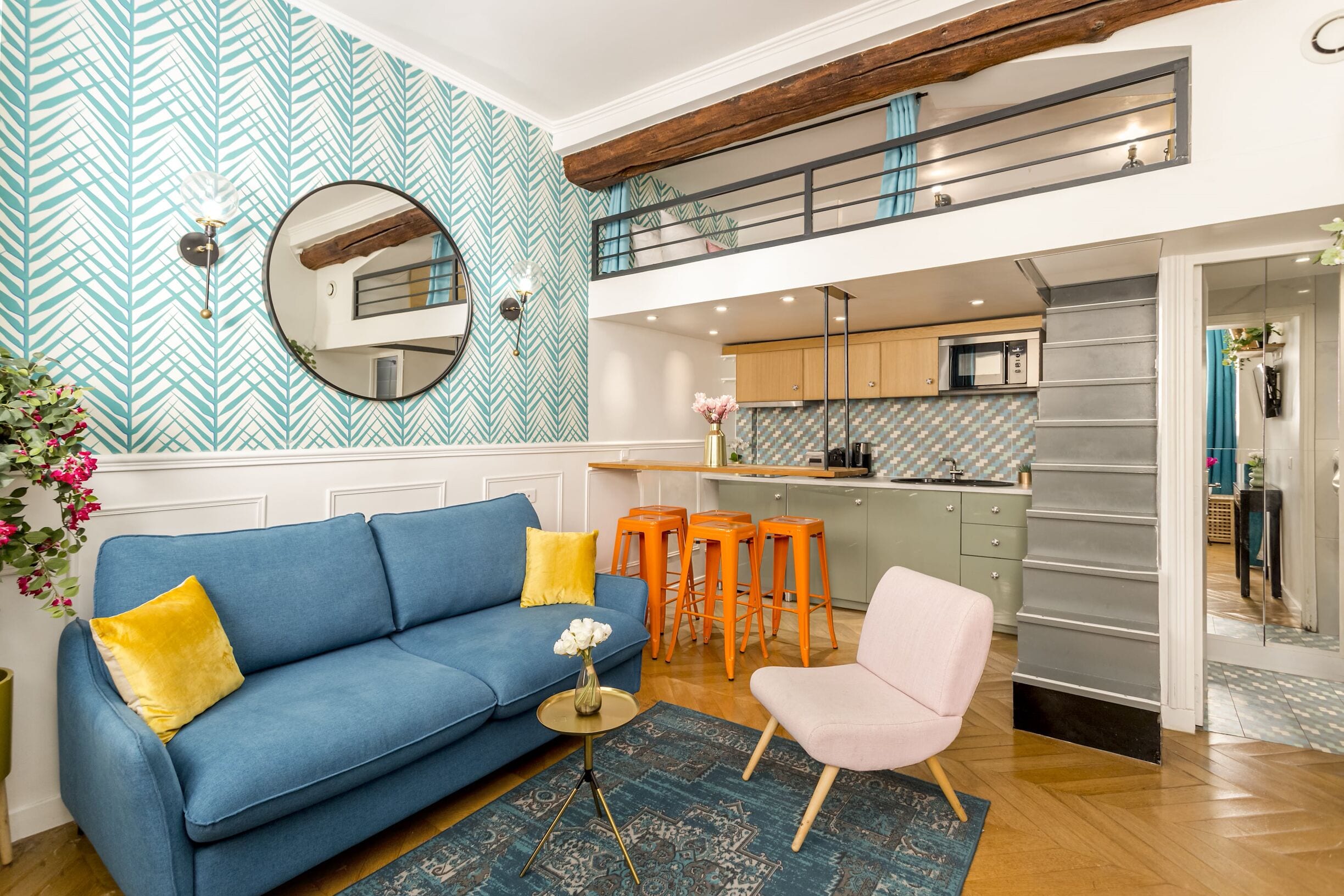 Un salon moderne avec un canapé bleu, un fauteuil rose et des tabourets orange. On aperçoit une cuisine à l’arrière-plan, avec un dosseret de crédence en carreaux disposés en motif chevrons.