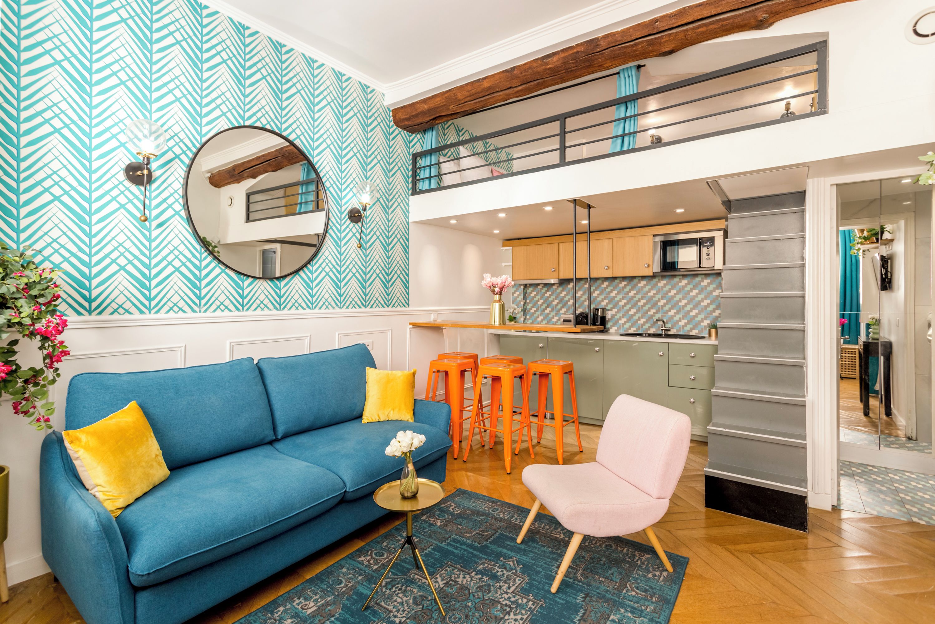 A modern living room with a blue sofa, a pink chair, and orange stools. A kitchen is visible in the background with a herringbone tile backsplash.