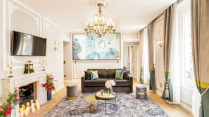 A living room with a fireplace, a sofa, a chandelier, and a large abstract painting. - Merveil - Signature Suite - Louvre - Victoria (Paris)