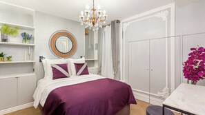 A bedroom with a bed, a wardrobe, a vanity with a mirror, a chandelier, and a table with a vase of flowers. - Merveil - Signature Suite - Louvre - Victoria (Paris)