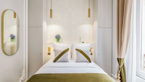Habitación de hotel con una cama grande, un espejo, una mesita de noche con un florero y una ventana con cortinas.