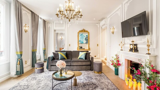 A living room with a grey sofa, a round coffee table, a chandelier, a fireplace, and a TV mounted on the wall. - Merveil - Signature Suite - Louvre - Victoria (Paris)