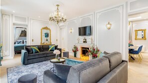 A spacious living room with a grey sofa, a blue armchair, and a coffee table. There is a fireplace with a clock above it, and a TV mounted on the wall. - Merveil - Signature Suite - Louvre - Victoria (Paris)