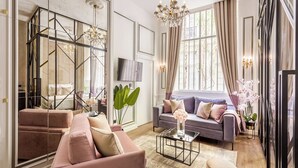 A modern living room with a sofa, a coffee table, and a TV. There is a large window with curtains and a chandelier. - Merveil - Luxury Suite - Marais - Archives I (Paris)