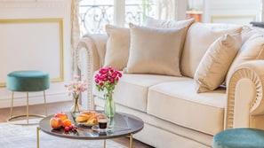 A living room with a beige sofa, a round coffee table with fruits, pastries, and flowers, and a teal ottoman. - Merveil - Luxury Serviced Apartment - Aboukir VI (Paris)