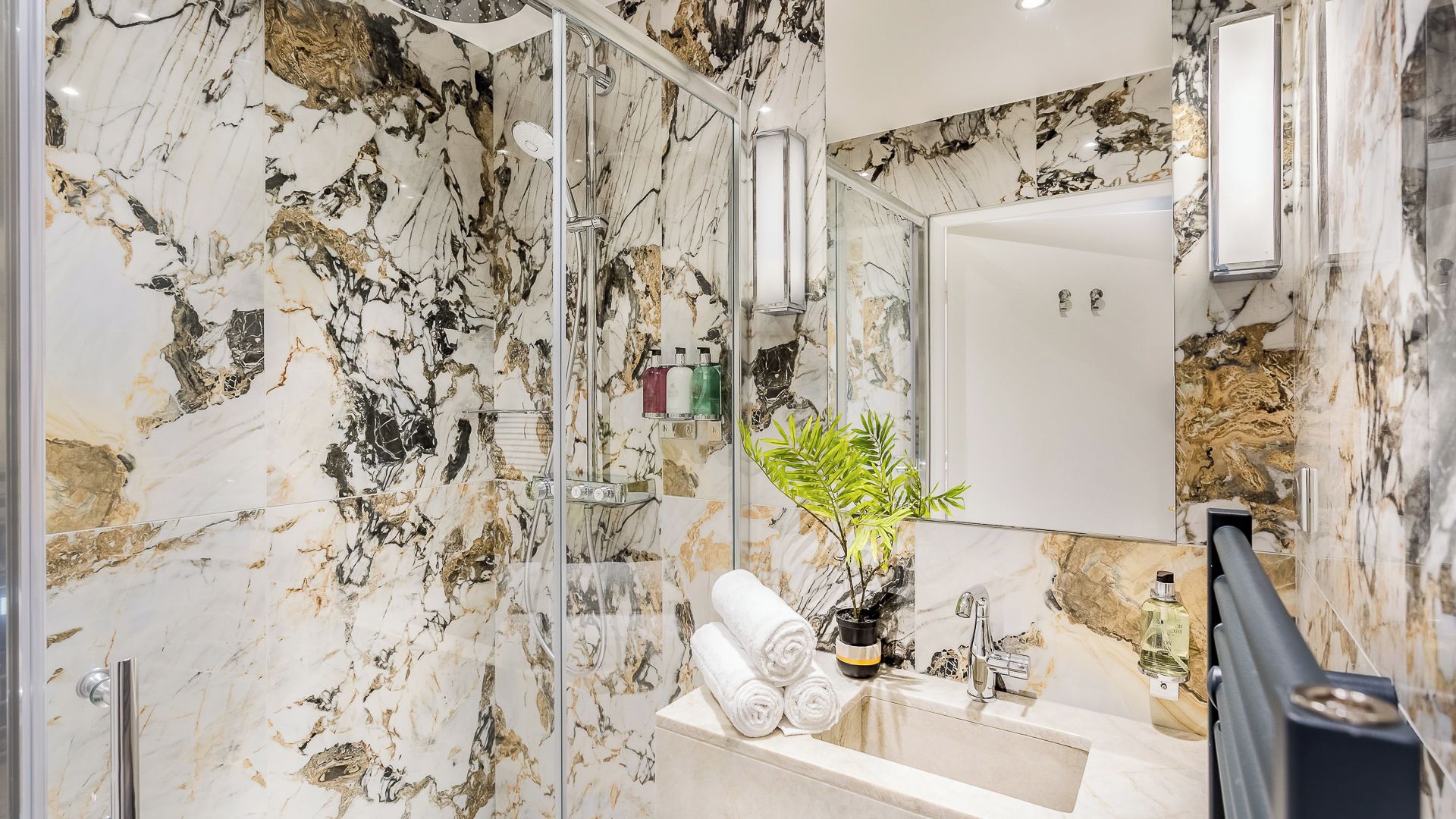 A bathroom with marble walls, a glass-enclosed shower, a sink with a mirror, and a potted plant.