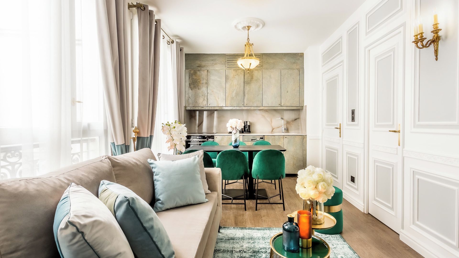 A modern living room with a sofa, green chairs, and a dining table. There is a kitchen area with cabinets and a chandelier.