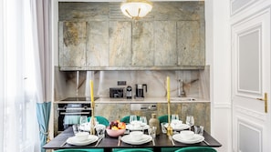 A modern kitchen with a stone backsplash, built-in appliances, and a dining area set for six with green chairs and a table set for a meal. - Merveil - Luxury Suite - Louvre - Marcel II (Paris)