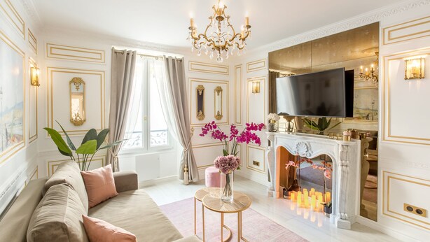 Una sala de estar con un sofá color beige, una alfombra rosa, una mesita redonda pequeña, una chimenea y un televisor.