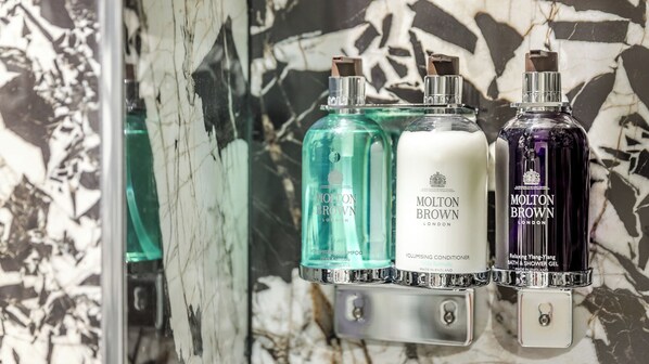 A bathroom with a marble wall and a mirror. There are three bottles of Molton Brown products: a volumising shampoo, a volumising conditioner, and a relaxing shower gel.