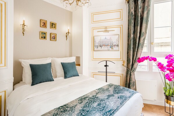 A well-appointed hotel room with a large bed, a chandelier, framed artwork, and a view of the city.