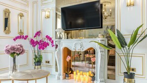 Una habitación lujosa con un televisor de pantalla plana grande, espejos ornamentados, una chimenea blanca con velas encendidas y plantas en macetas.