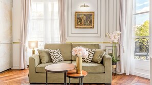 A living room with a sofa, a round table, a vase with flowers, a painting on the wall, and a view of the outdoors. - Merveil - Luxury Suite - Louvre -Sebastopol I (Paris)
