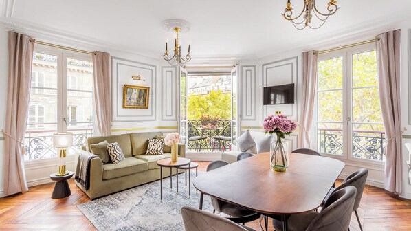 A spacious living room with a sofa, a dining table, and chairs, a chandelier, and a balcony with a view.