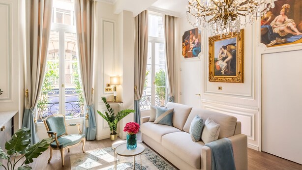 Un salon bien éclairé, agrémenté d’un lustre, d’un canapé, d’un fauteuil et d’un vase de fleurs.