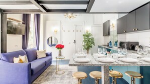 A modern living room with a blue sofa, a small round table with a vase of red flowers, and a dining area with a marble countertop and bar stools.