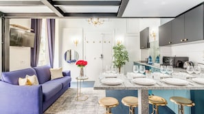 Ein modernes Wohnzimmer mit einem blauen Sofa, einem kleinen runden Tisch mit einer Vase roter Blumen und einem Essbereich mit Marmorplatte und Barhocker.