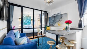 Habitació d'hotel moderna amb un sofà blau, una zona de menjador amb un respatller de marbre i una finestra gran amb cortines.