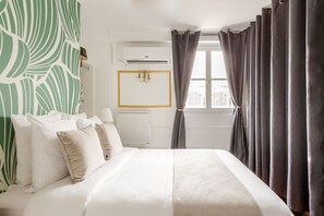 A neatly made bed with white and beige bedding, a wall-mounted air conditioner, and a window with curtains.
