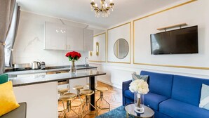 Une cuisine moderne avec des meubles blancs, un plan de travail en marbre et un îlot central doté de tabourets. On y trouve un canapé bleu orné de motifs floraux blancs et un téléviseur fixé au mur.