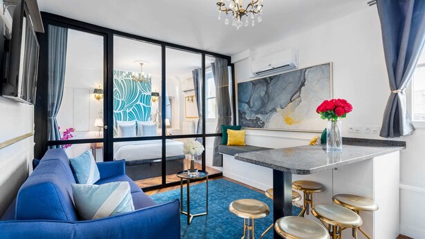 Une chambre d’hôtel moderne avec un canapé bleu, un coin repas doté d’un pare-feu en marbre et une grande fenêtre avec des rideaux.