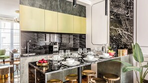 A modern kitchen with a marble backsplash, a dining area with a long table set for six, and a bar area with stools. - Merveil - Luxury Suite - Louvre - Sebastopol II (Paris)