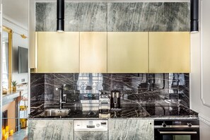 A modern kitchen with a black marble backsplash, stainless steel appliances, and gold-toned cabinets. - Merveil - Luxury Suite - Louvre - Sebastopol II (Paris)