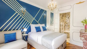 A modern bedroom with a geometric blue and gold wall design, a white bed with blue pillows, a beige sofa, a chandelier, and a marble-patterned wall. - Merveil - Luxury Suite - Louvre - Sebastopol II (Paris)