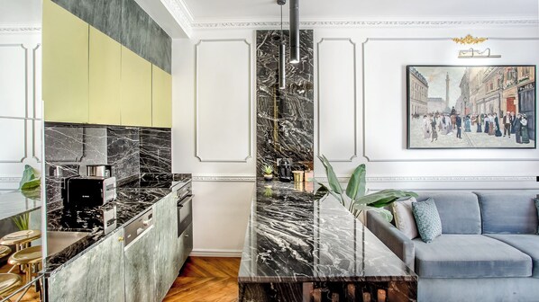 A modern kitchen with a black marble countertop, a Bosch coffee machine, and a grey sofa in the living area. - Merveil - Luxury Suite - Louvre - Sebastopol II (Paris)