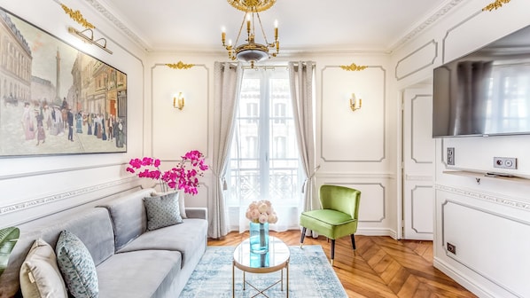 A well-lit living room with a sofa, a green armchair, a TV, and a painting of a street scene. - Merveil - Luxury Suite - Louvre - Sebastopol II (Paris)