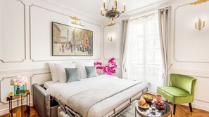 A hotel room with a large bed, a green armchair, a small table with breakfast, and a painting of a city scene. - Merveil - Luxury Suite - Louvre - Sebastopol II (Paris)