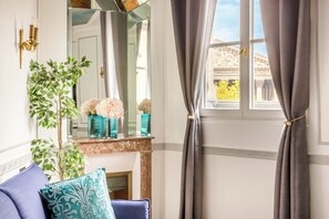 A room with a large mirror, a potted plant, a sofa with patterned cushions, and a window with curtains. - Merveil Luxury Suite Royale II - Saint Honore view (Paris)