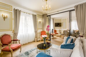 Un salon luxueux agrémenté d’un lustre, de fauteuils rouges et dorés, d’un canapé avec des coussins bleus, et d’un coin repas à l’arrière-plan.