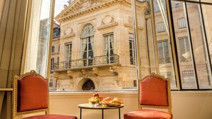A room with two red upholstered chairs, a small round table with a fruit bowl, a glass of juice, and a plate of pastries. A large window with a balcony overlooking a historic building is visible. - Merveil Signature Suite - Valois - Louvre view (Paris)