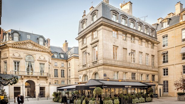 A historic building with classical architecture, featuring a central courtyard with outdoor seating and a covered area. - Merveil Signature Suite - Valois - Louvre view (Paris)