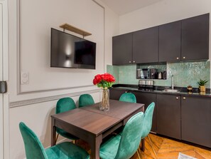 A modern kitchen with dark cabinets, a flat-screen TV, and a dining area with teal chairs and a wooden table. - Merveil - Luxury Suite - Louvre - Aboukir IV (Paris)