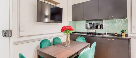 Una cocina moderna con armarios oscuros, un televisor de pantalla plana montado sobre la encimera y una zona de comedor con una mesa de madera y sillas color turquesa.
