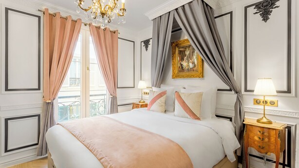 Ein Hotelzimmer mit einem großen Bett, zwei Nachttischen mit Lampen, einer Chandelüster und einem Balkon mit Vorhängen.