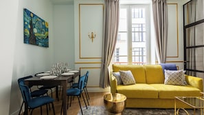 Zona pranzo con tavolo apparecchiato per quattro persone, un divano giallo con cuscini decorati e un quadro appeso alla parete.