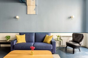 Een moderne woonkamer met een blauwe bank, gele kussens, een houten salontafel en een zwarte fauteuil.