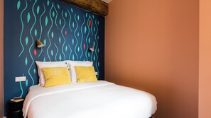 Un lit avec une literie blanche et deux oreillers jaunes, un mur orné d’un motif abstrait aux couleurs turquoise et rouge, et une seule lampe de chevet.