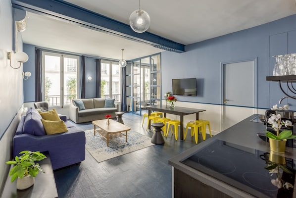 Een moderne woonkamer met een bank, salontafel en gele krukken. Er is een keuken met een fornuis en een eettafel.