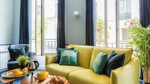 Een woonkamer met een gele bank, donkere gordijnen, een wandlamp en uitzicht op de straat door grote ramen.