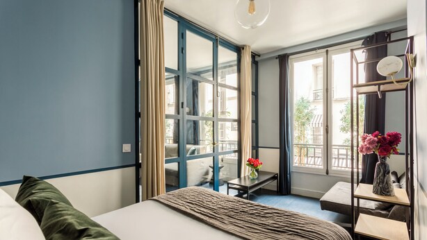 Habitación de hotel con cama, una mesita, una silla, una estantería con flores y vistas al exterior.