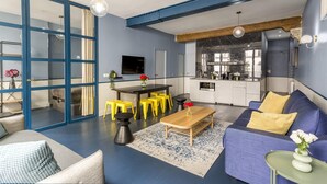 Una sala de estar moderna con un sofá azul, sillones amarillos, una mesa de centro de madera y una cocina al fondo.