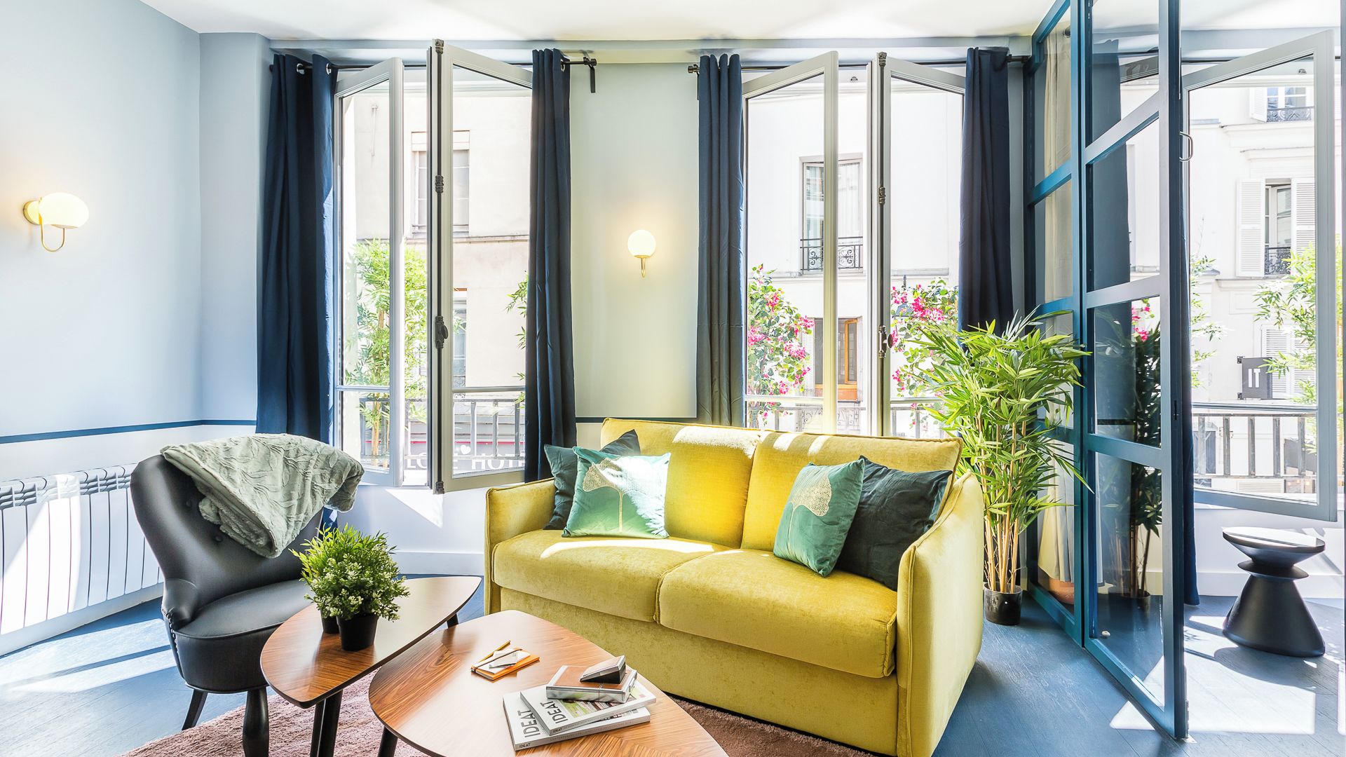 A modern living room with a yellow sofa, black armchair, and a small coffee table. Large windows with curtains provide views of the street.