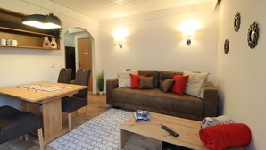 Ein Wohnzimmer mit einem Holzespeisetisch, einem braunen Sofa mit roten und weißen Kissen, einem Couchtisch mit Fernbedienung und einem roten Handtuch sowie einem gemusterten Teppich.