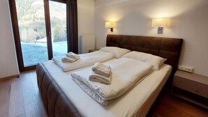 Ein Doppelbett mit weißer Bettwäsche und Handtüchern, ein hölzernes Kopfteil, zwei Nachttischlampen und ein Blick auf eine gebirgige Landschaft durch ein großes Fenster.