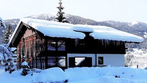 Ein Chalet mit schneebedecktem Dach und großen Fenstern, umgeben von schneebedeckten Bäumen und Bergen.